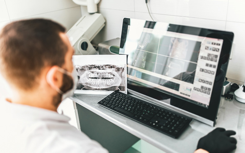 A man wearing a white lab coat is sitting at a desk with two laptops open in front of him, working on one while looking at another, with a digital rendering displayed on the screen.